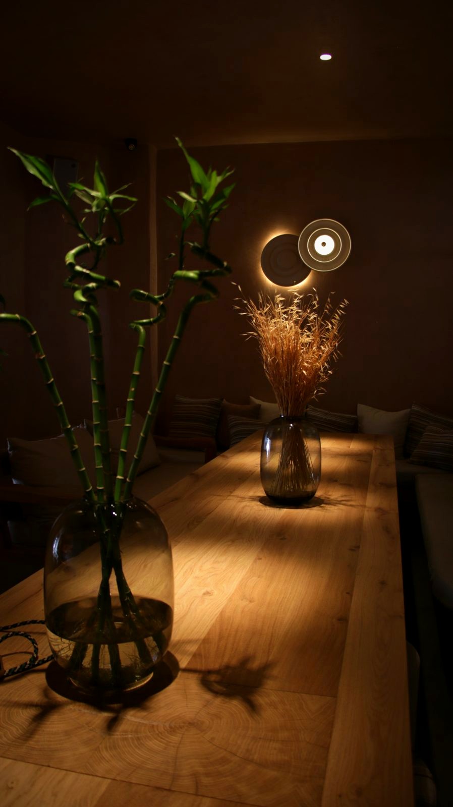 A long timber table set with glass vases at Bouboulo, lit warm by round wall sconces in a dark room.