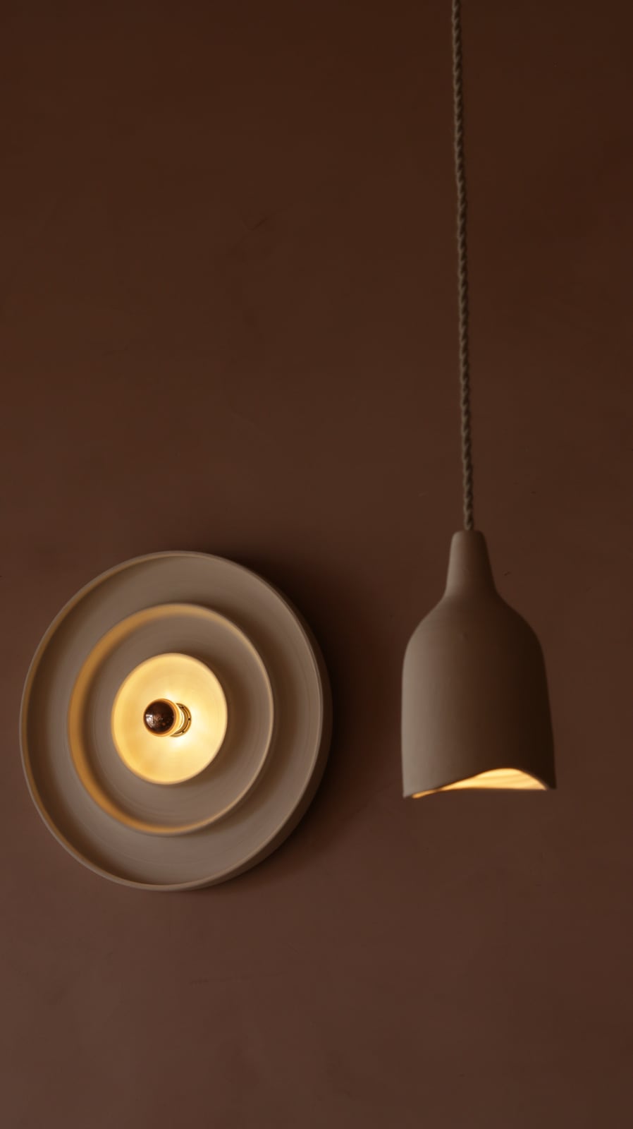 A round ceramic sconce and a ceramic pendant glowing side by side on a clay colored wall at Bouboulo.