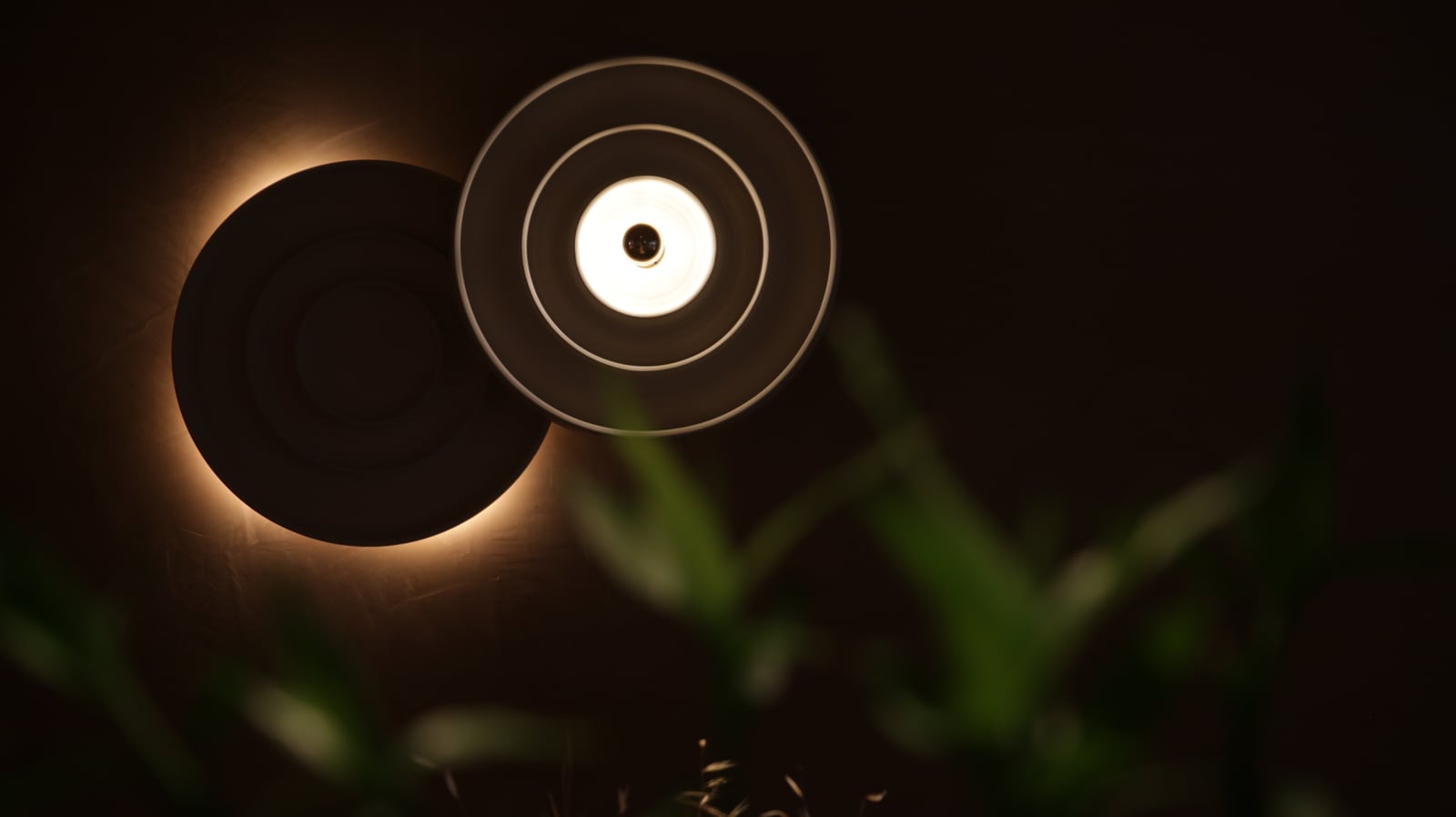 Two round sconces on a dark wall at Bouboulo, one a dark disc ringed by its own halo, the other a bright ring, leaves blurred in front.