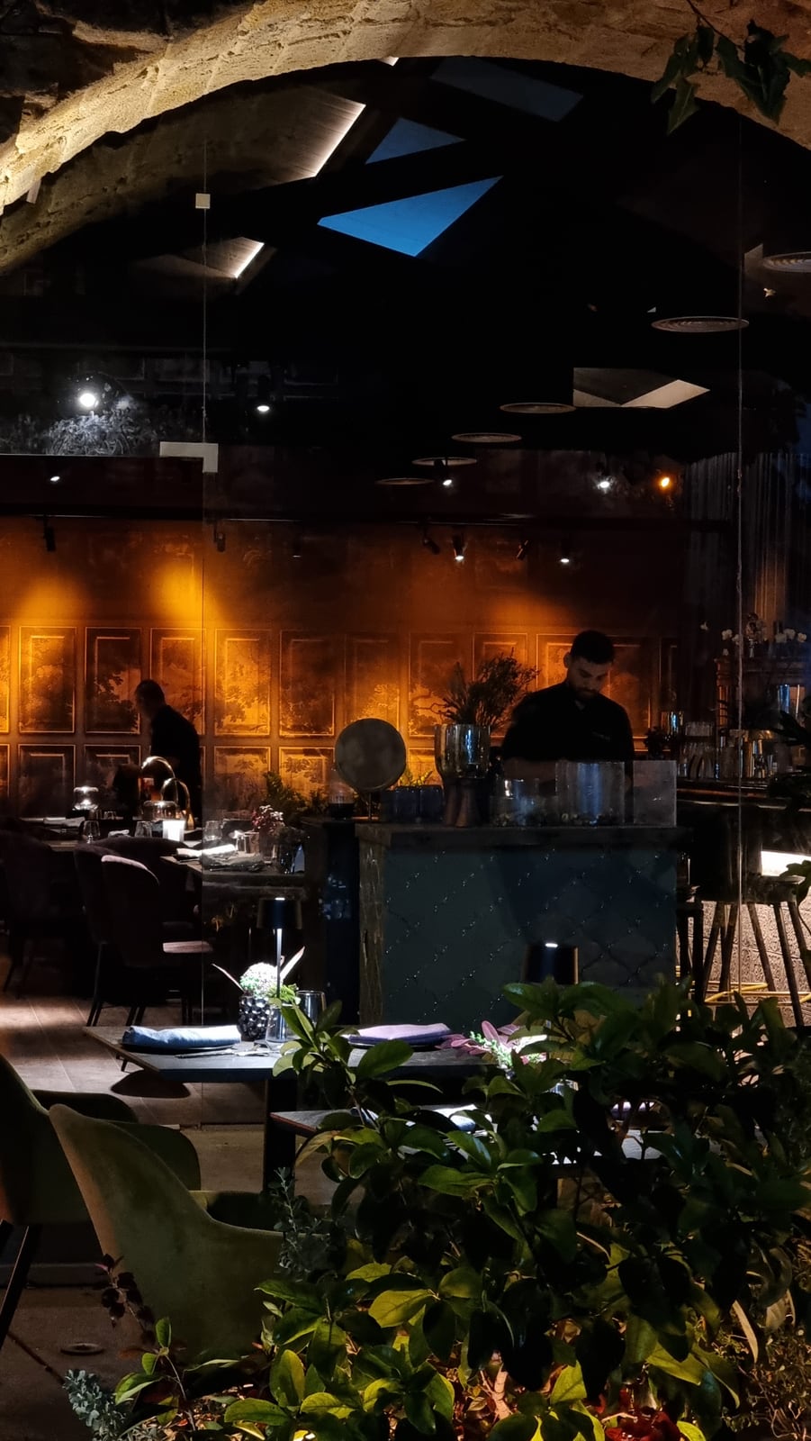 A stone vault opening onto blue skylights above the Itameshi dining room, warm light grazing a wall of painted panels behind the counter.