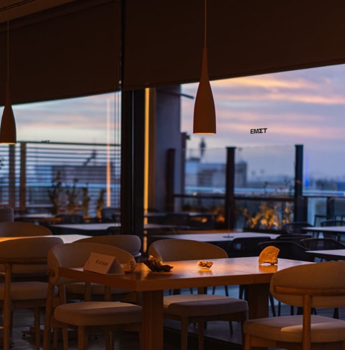 Slender pendants over timber tables in the Nyn Esti dining room at dusk, the city and a lilac sky beyond the glass.