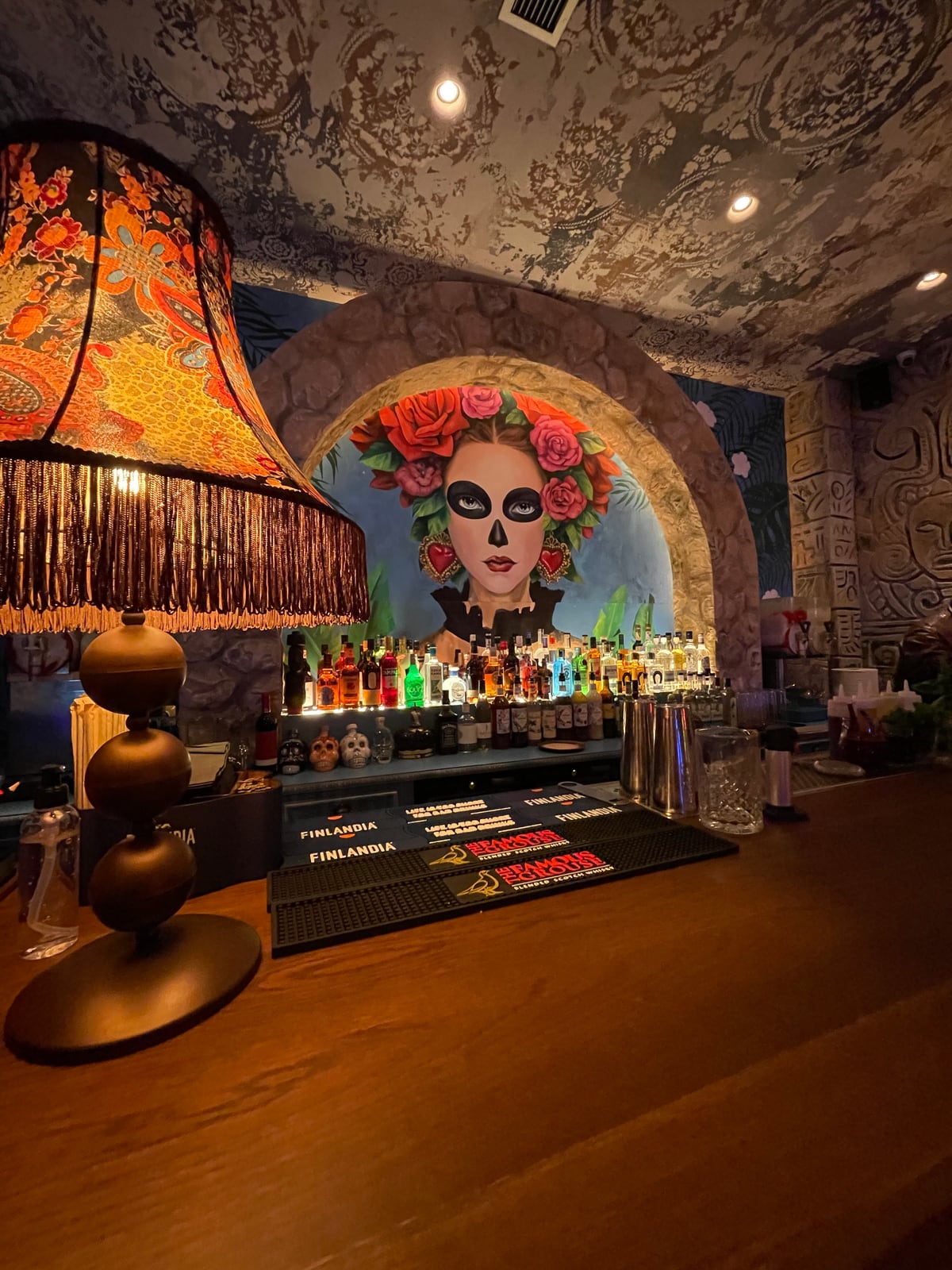 A fringed, patterned lampshade glowing on the Panchito bar, backlit bottles beneath a painted portrait in an arch.