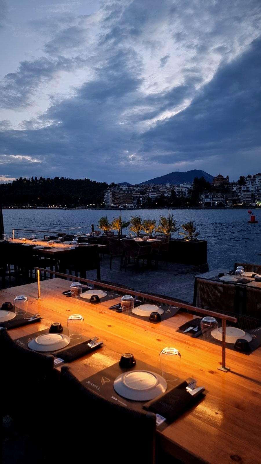 Long timber tables set for dinner on the Raval terrace, the bay and the hills beyond at dusk.