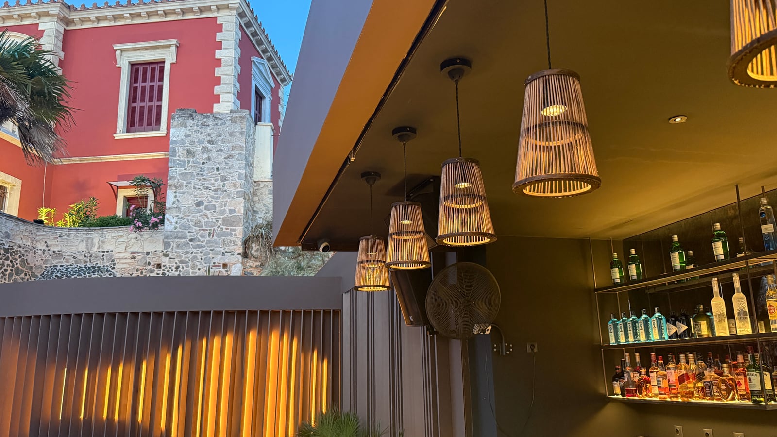Rattan pendants glowing under the Raval bar canopy at dusk, shelves of bottles lit behind and warm light between the slats of the fence, a red villa beyond.
