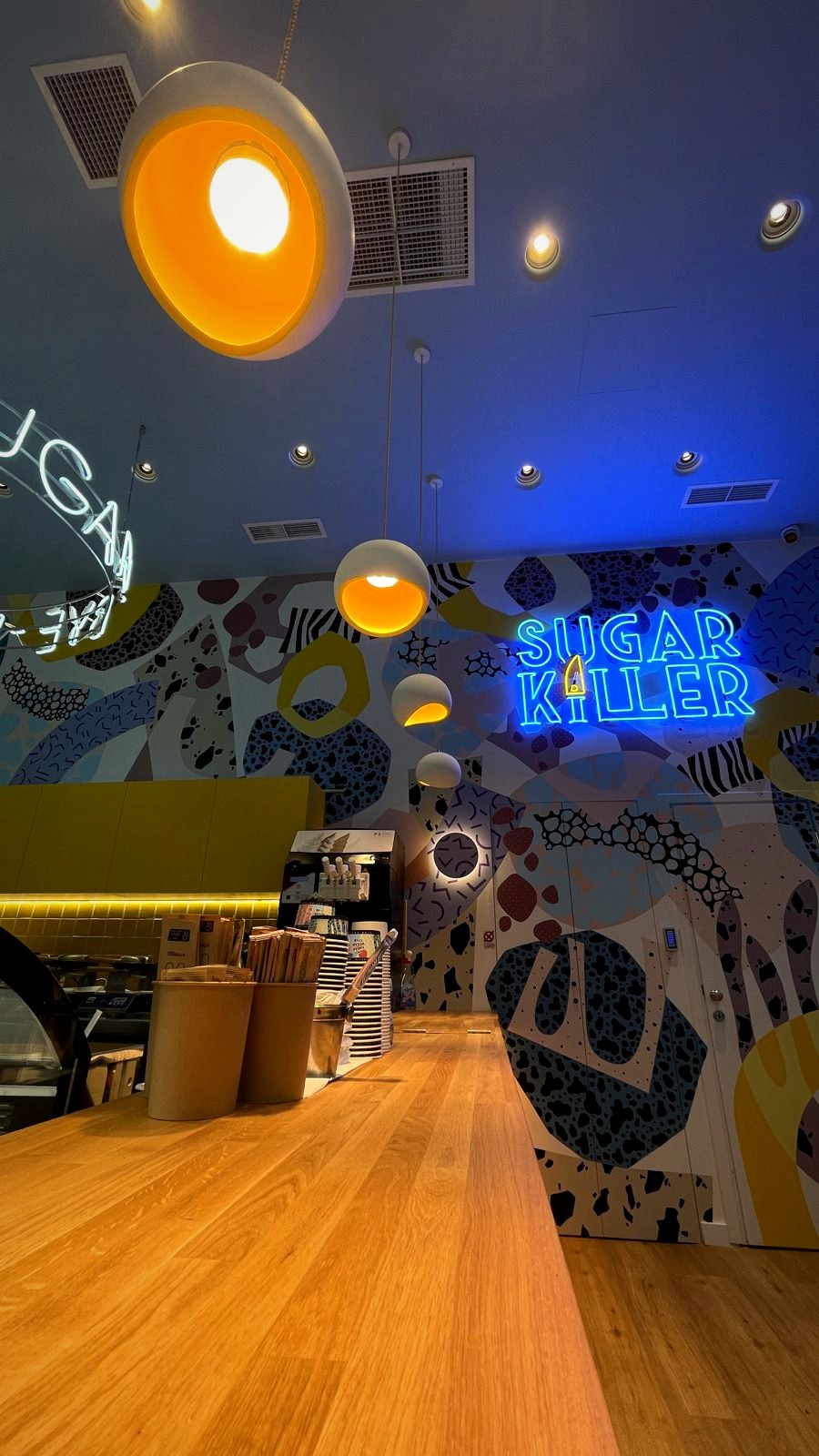 Sugar café's long timber counter beneath orange pendants, a patterned mural and a blue neon sign.