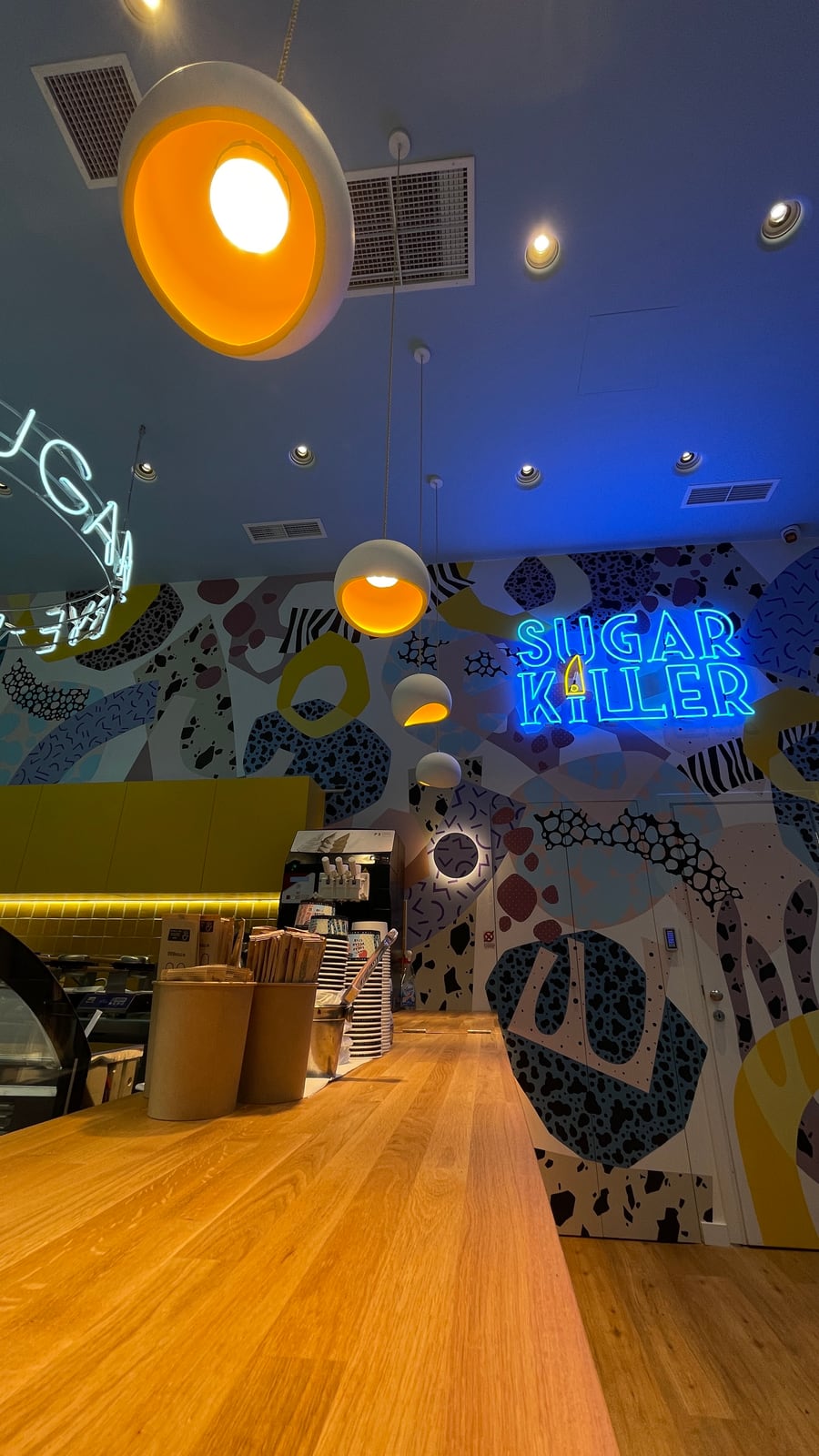 Orange lit ceramic pendants before a patterned mural and a blue neon sign at Sugar café, a round wall light casting a halo below.