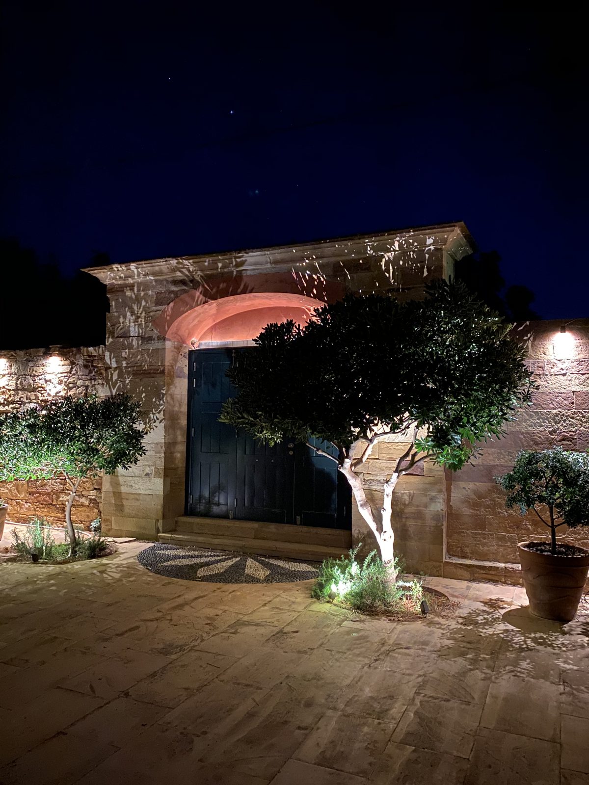 Olive trees and potted plants lit from below beside the villa's stone gateway.